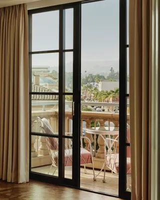 Balcony scene framed by black‑steel doors: pink‑cushioned chairs and round table behind a stone balustrade, overlooking palm‑lined Beverly Hills streets.