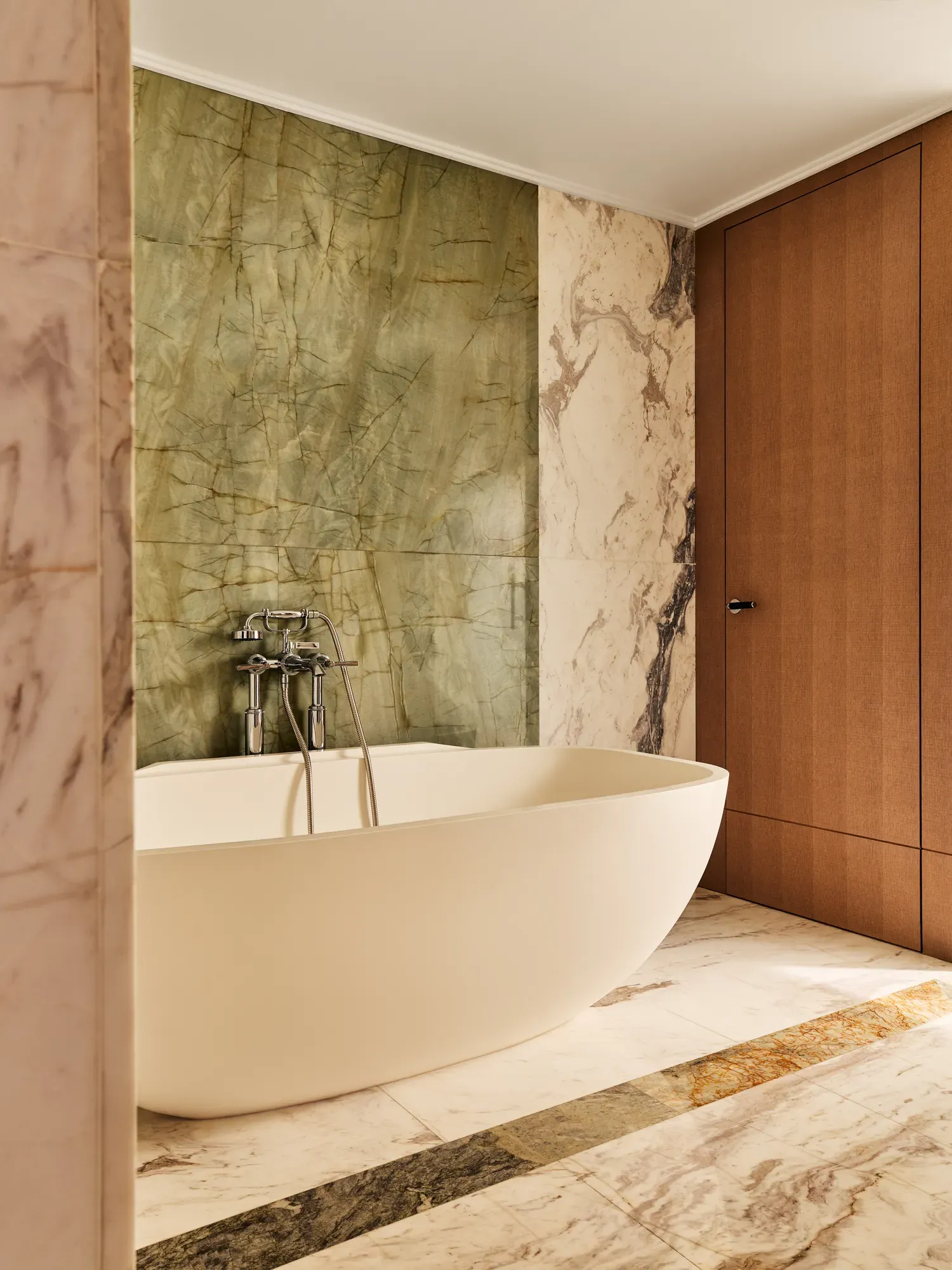 Elegant bathroom with freestanding oval bathtub, marble flooring, and green onyx accent wall beside wooden door.