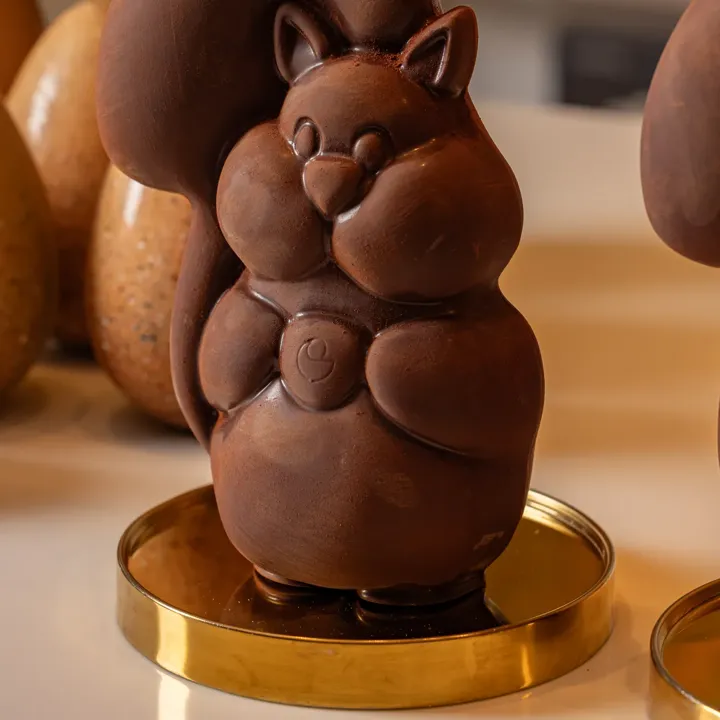 Chocolate squirrel Easter figurine on a gold stand with speckled chocolate eggs blurred in the background.