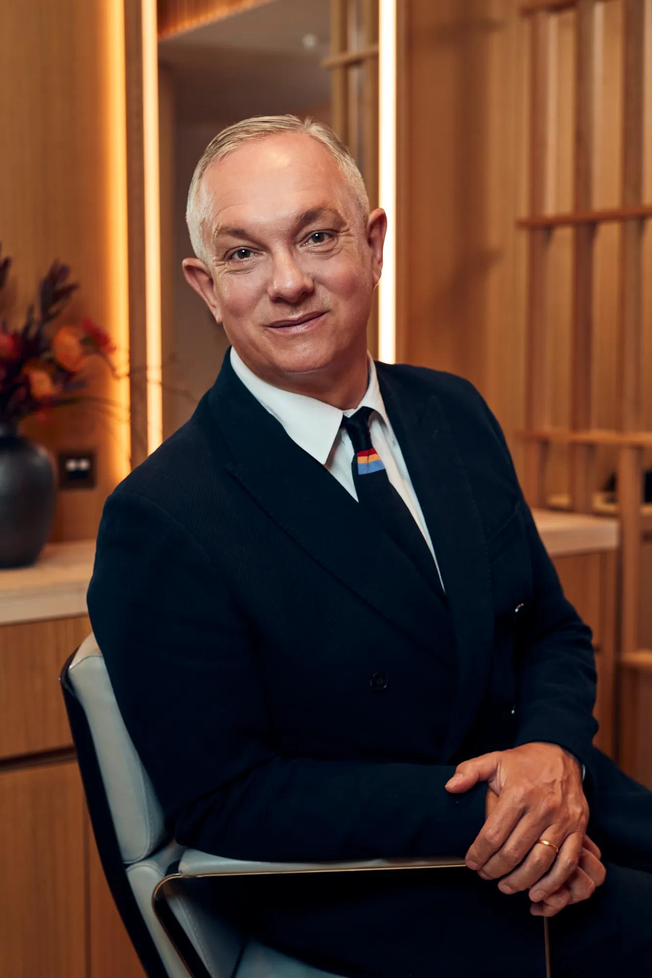 A man in a dark suit and tie sits in a modern wood-panelled room at The Maybourne Riviera, smiling warmly toward the camera.