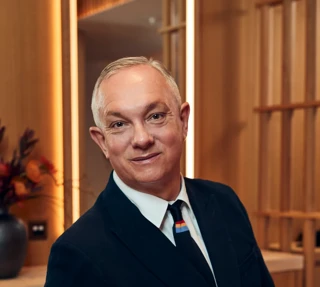 A man in a dark suit and tie sits in a modern wood-panelled room at The Maybourne Riviera, smiling warmly toward the camera.