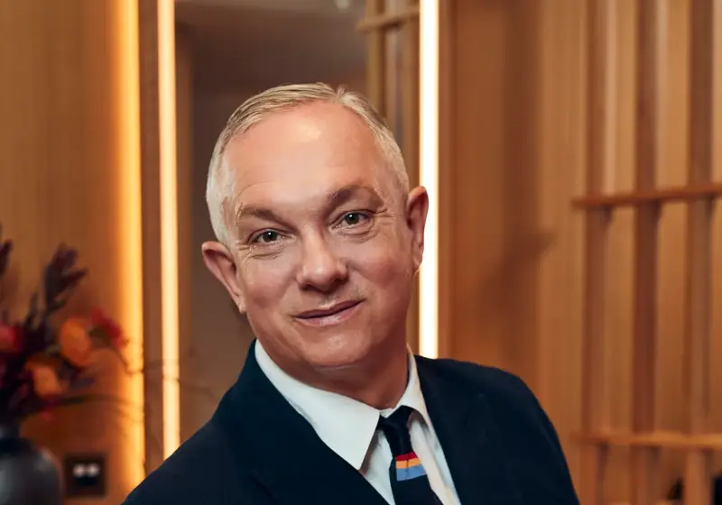 A man in a dark suit and tie sits in a modern wood-panelled room at The Maybourne Riviera, smiling warmly toward the camera.