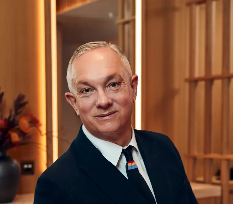 A man in a dark suit and tie sits in a modern wood-panelled room at The Maybourne Riviera, smiling warmly toward the camera.