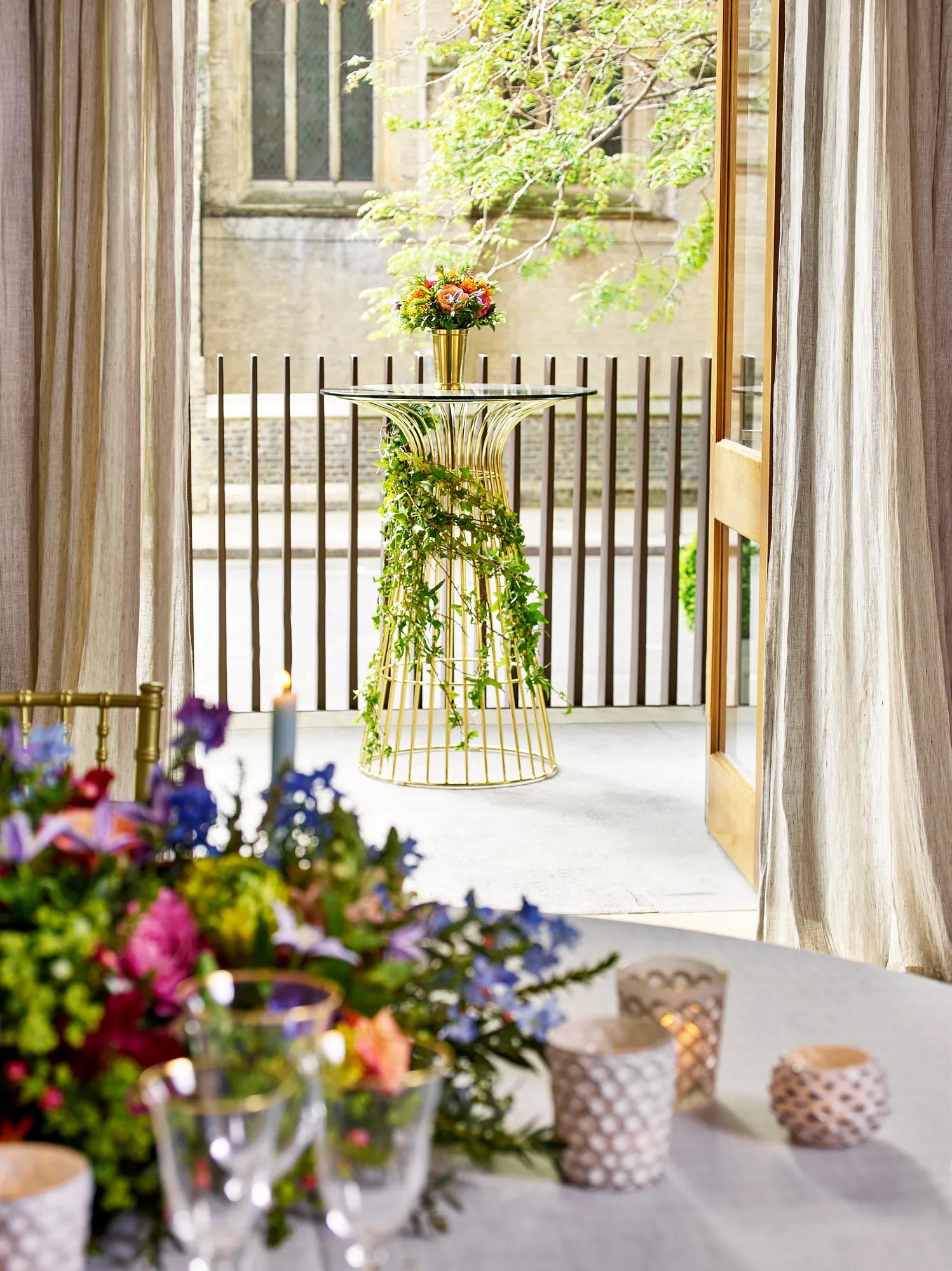 Gold cocktail table with ivy garland and flowers, placed outside near a terrace with draped curtains