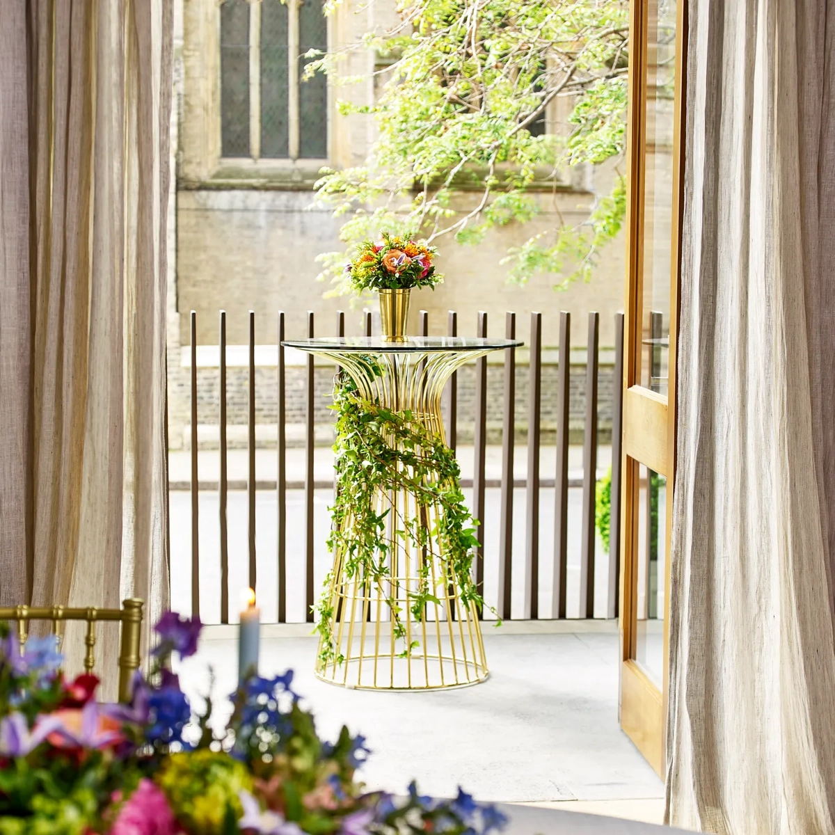 Gold cocktail table with ivy garland and flowers, placed outside near a terrace with draped curtains
