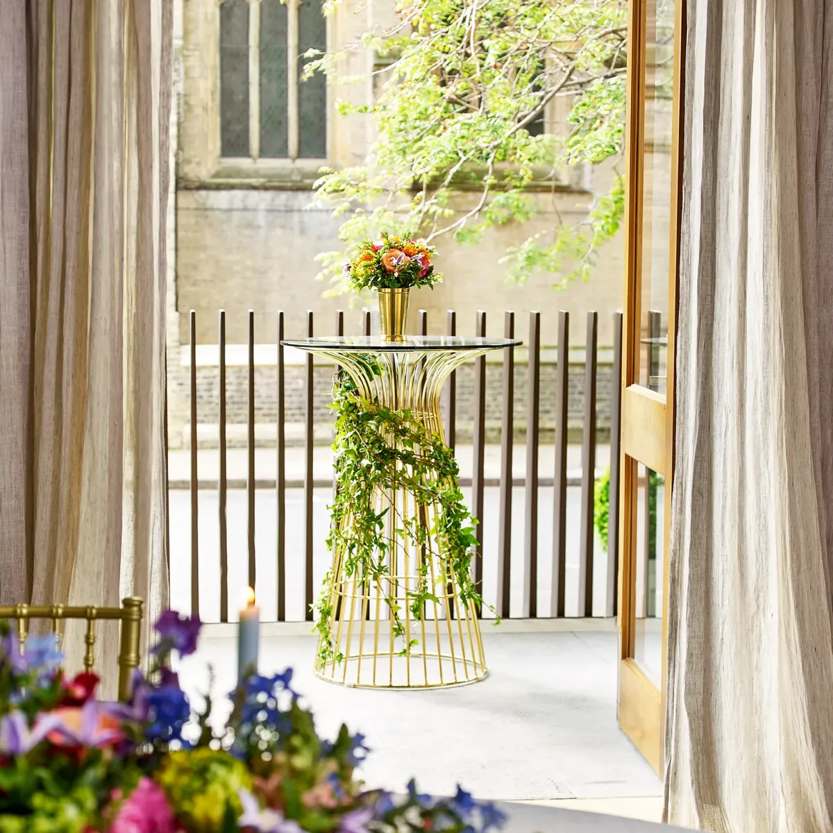 Gold cocktail table with ivy garland and flowers, placed outside near a terrace with draped curtains