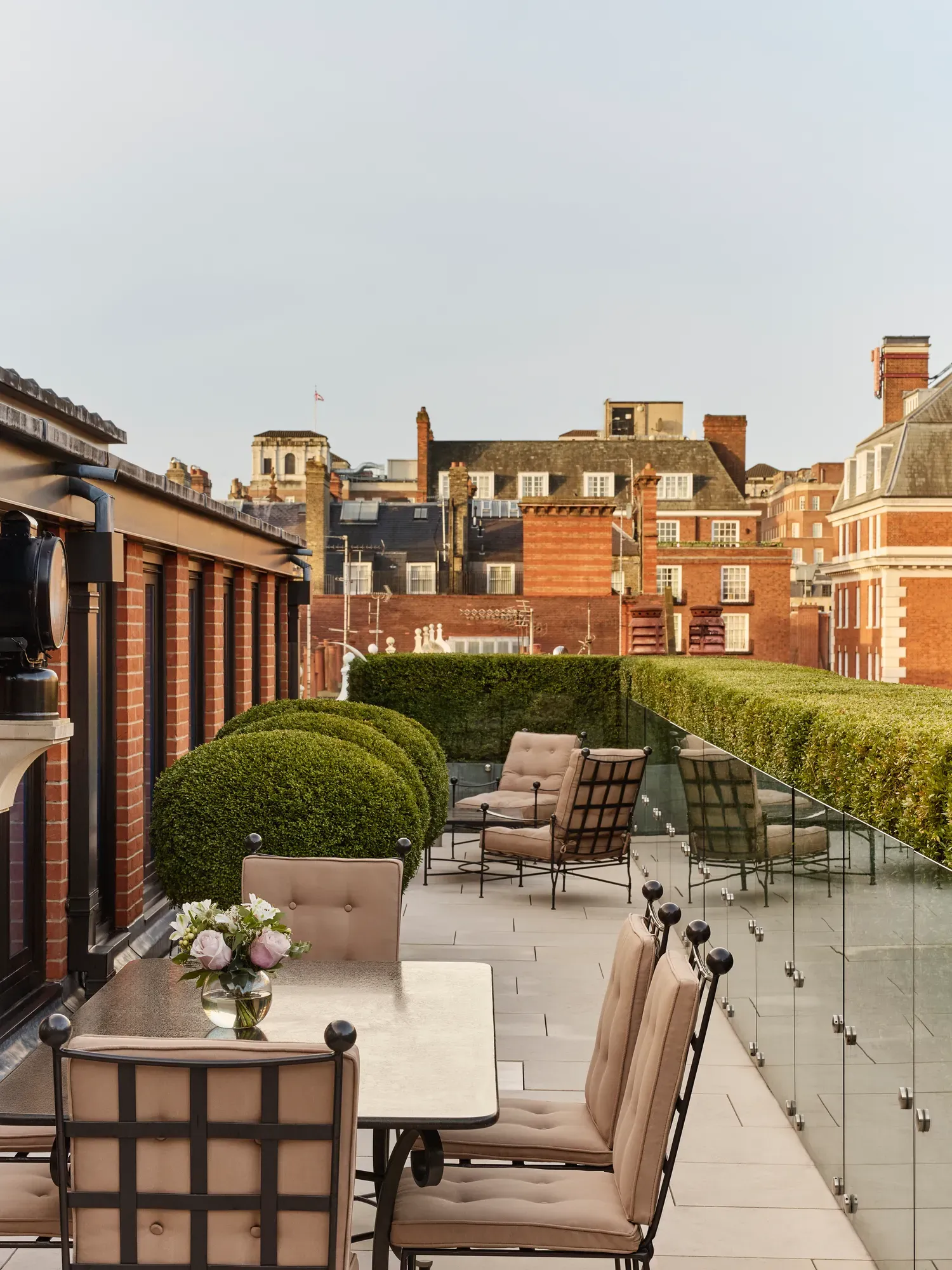 Terrace with cushioned dining chairs, lounge seating, manicured hedges, and views of red-brick rooftops.