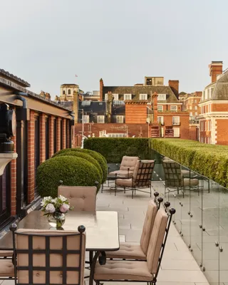 Terrace with cushioned dining chairs, lounge seating, manicured hedges, and views of red-brick rooftops.