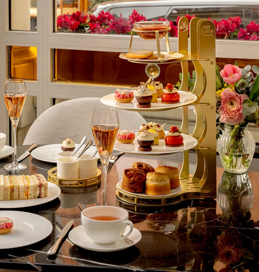 Afternoon tea table with tiered pastries, finger sandwiches, teacups, and glasses of rosé beside a vase of flowers.