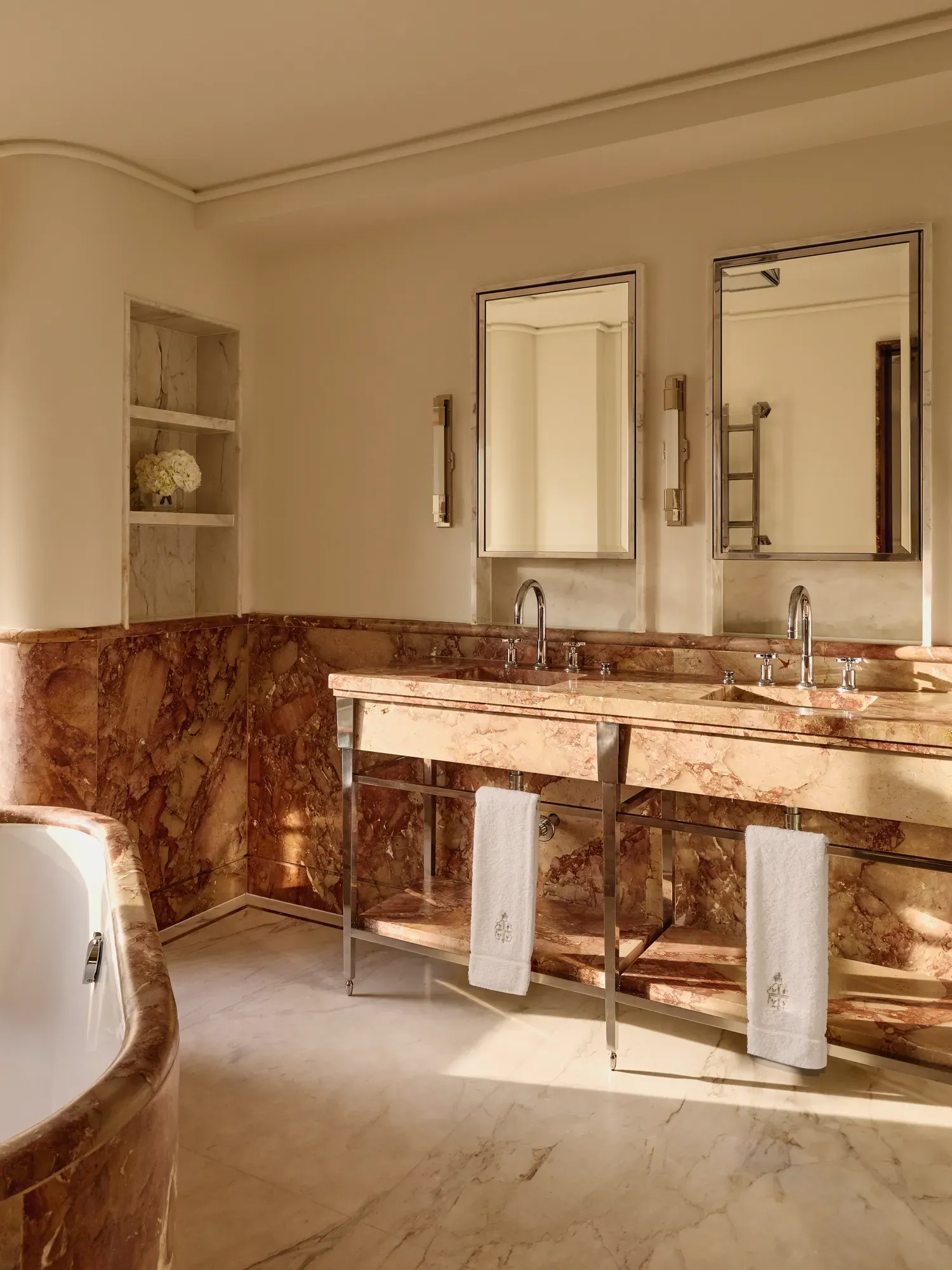 Elegant bathroom with rose-toned marble walls and double vanity, twin mirrors, chrome fittings, and soft natural light illuminating a corner bathtub.