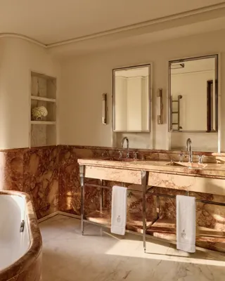Elegant bathroom with rose-toned marble walls and double vanity, twin mirrors, chrome fittings, and soft natural light illuminating a corner bathtub.
