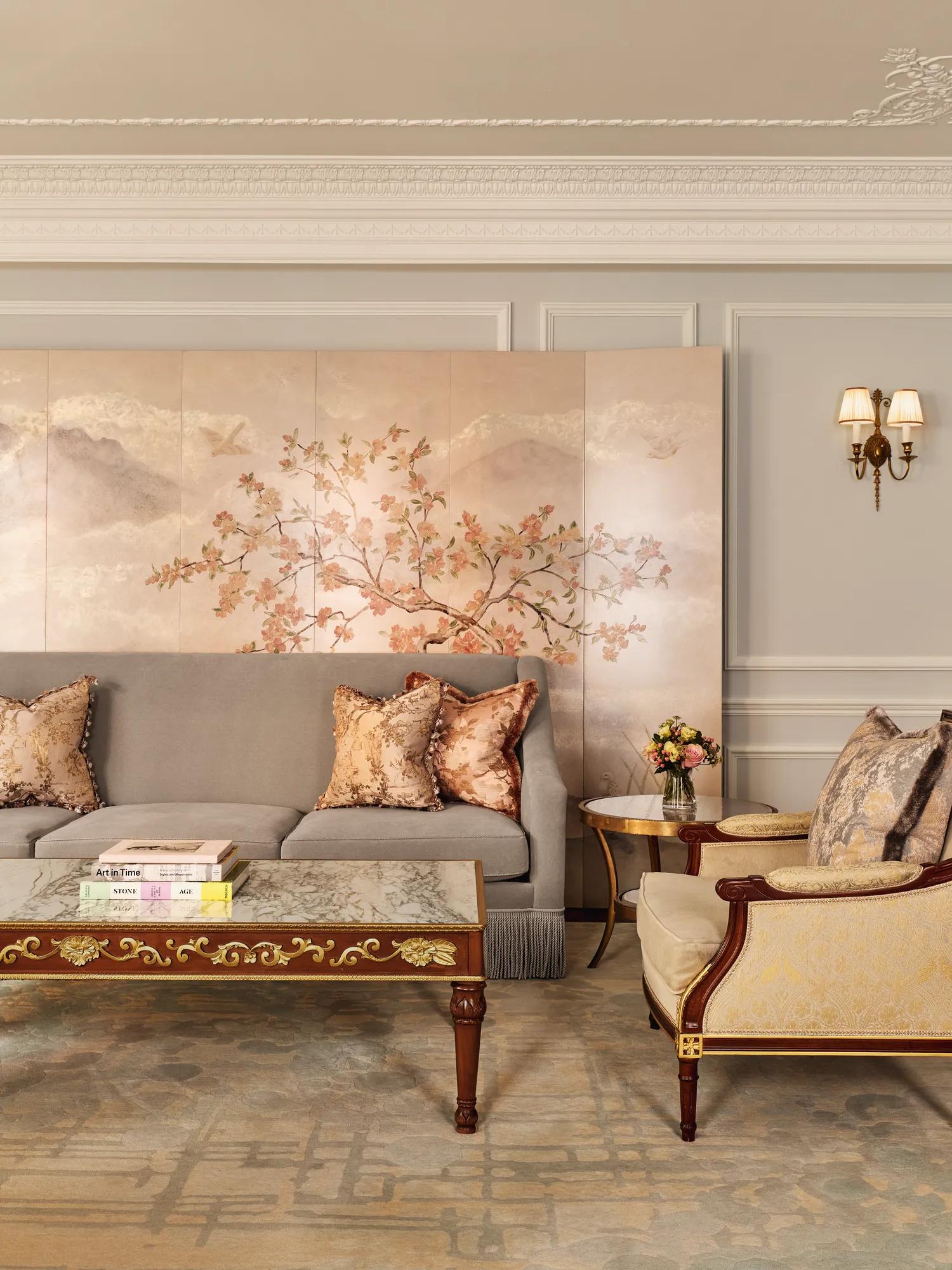 An elegant living room with a grey sofa and gold-patterned armchair, featuring silk cushions, a decorative coffee table with books, and a hand-painted wall panel of delicate branches and birds in soft tones.