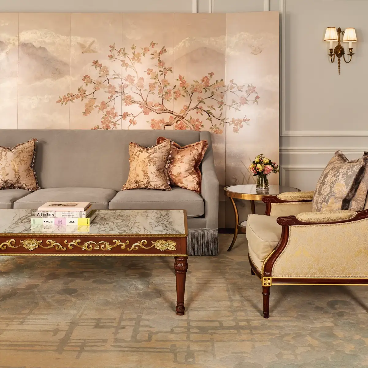 An elegant living room with a grey sofa and gold-patterned armchair, featuring silk cushions, a decorative coffee table with books, and a hand-painted wall panel of delicate branches and birds in soft tones.