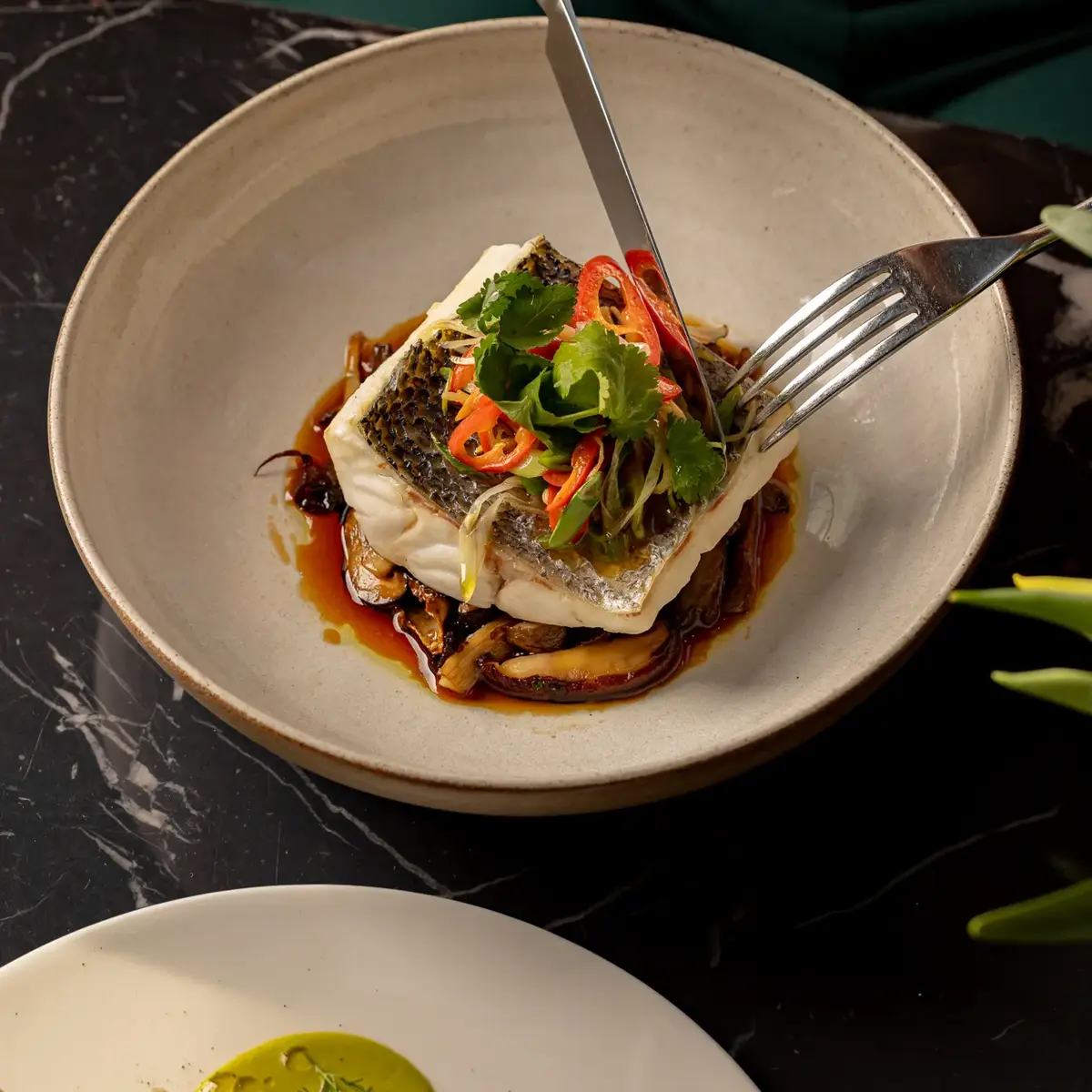 Person cutting into seared fish topped with herbs and sliced chili on a ceramic plate with a rich sauce.