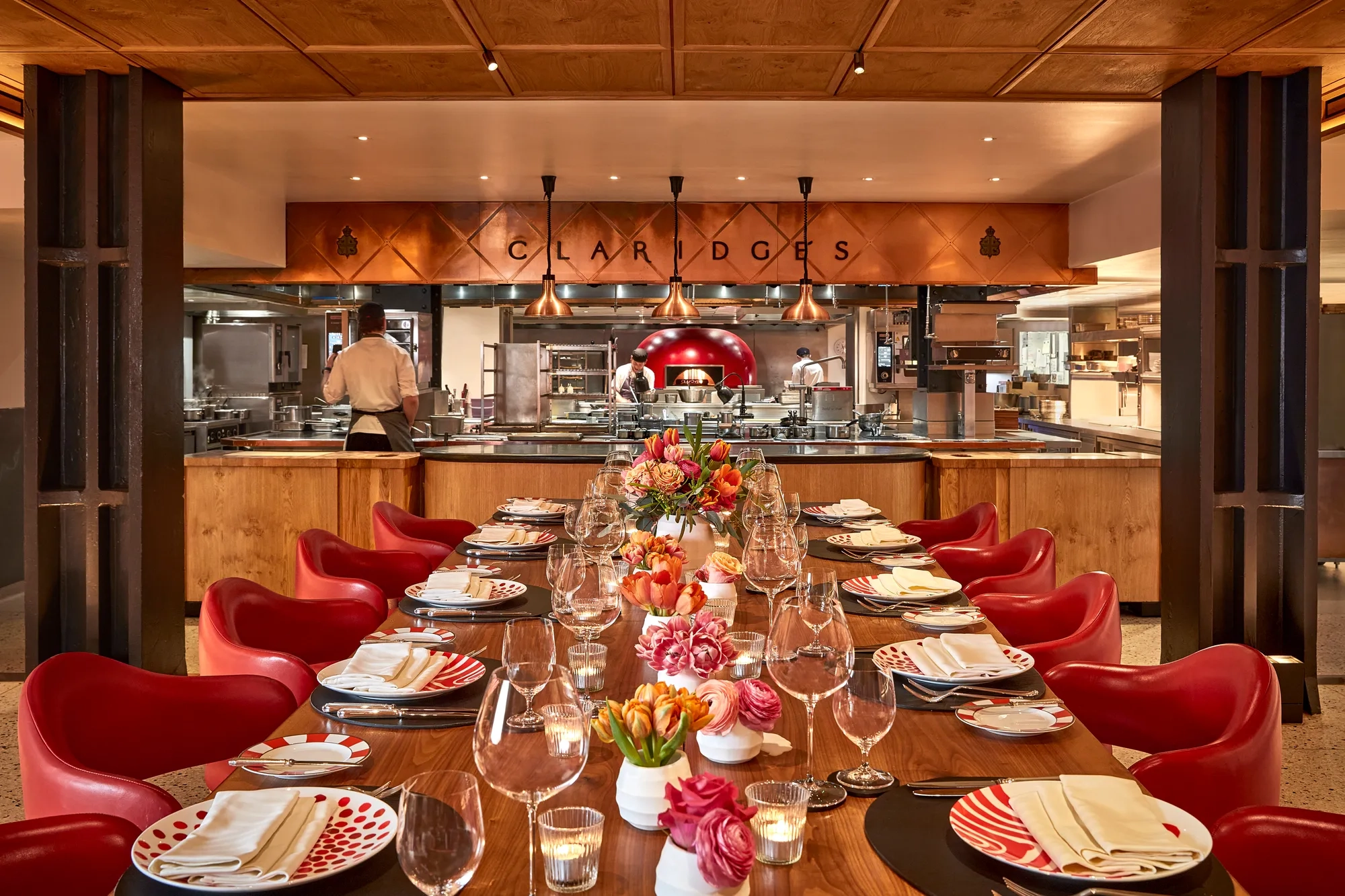 Elegant open-kitchen dining space at Claridge’s, featuring a long wooden table with red chairs, patterned plates, and floral centerpieces. The chefs work in the background beneath copper lighting, creating a warm and inviting atmosphere.