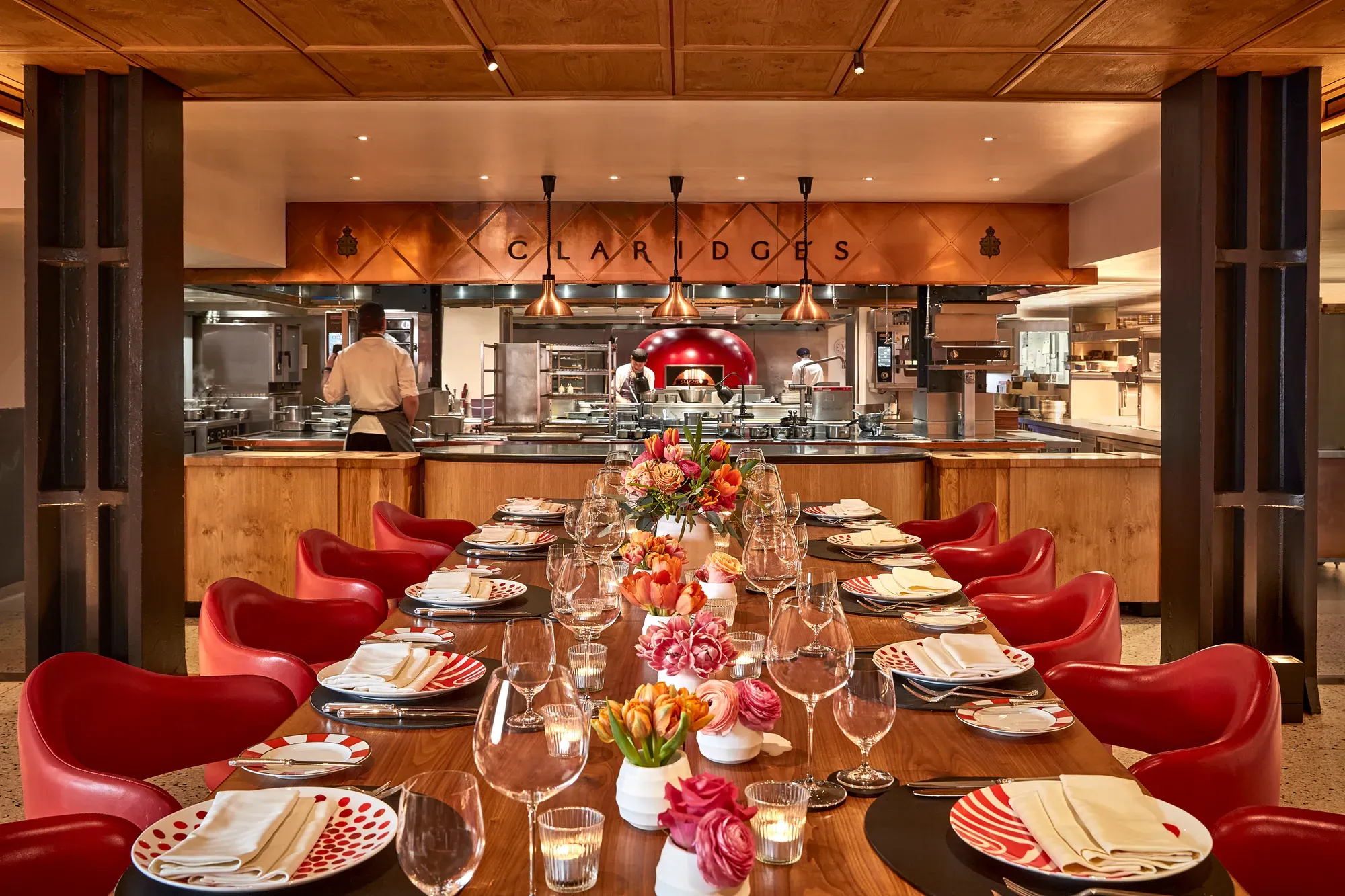 Elegant open-kitchen dining space at Claridge’s, featuring a long wooden table with red chairs, patterned plates, and floral centerpieces. The chefs work in the background beneath copper lighting, creating a warm and inviting atmosphere.