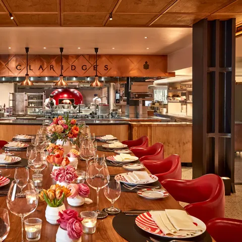 Elegant open-kitchen dining space at Claridge’s, featuring a long wooden table with red chairs, patterned plates, and floral centerpieces. The chefs work in the background beneath copper lighting, creating a warm and inviting atmosphere.