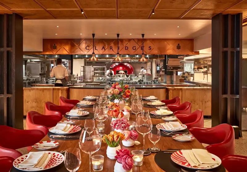 Elegant open-kitchen dining space at Claridge’s, featuring a long wooden table with red chairs, patterned plates, and floral centerpieces. The chefs work in the background beneath copper lighting, creating a warm and inviting atmosphere.
