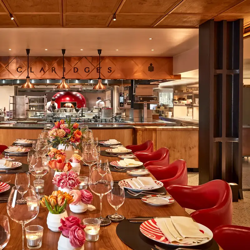 Elegant open-kitchen dining space at Claridge’s, featuring a long wooden table with red chairs, patterned plates, and floral centerpieces. The chefs work in the background beneath copper lighting, creating a warm and inviting atmosphere.