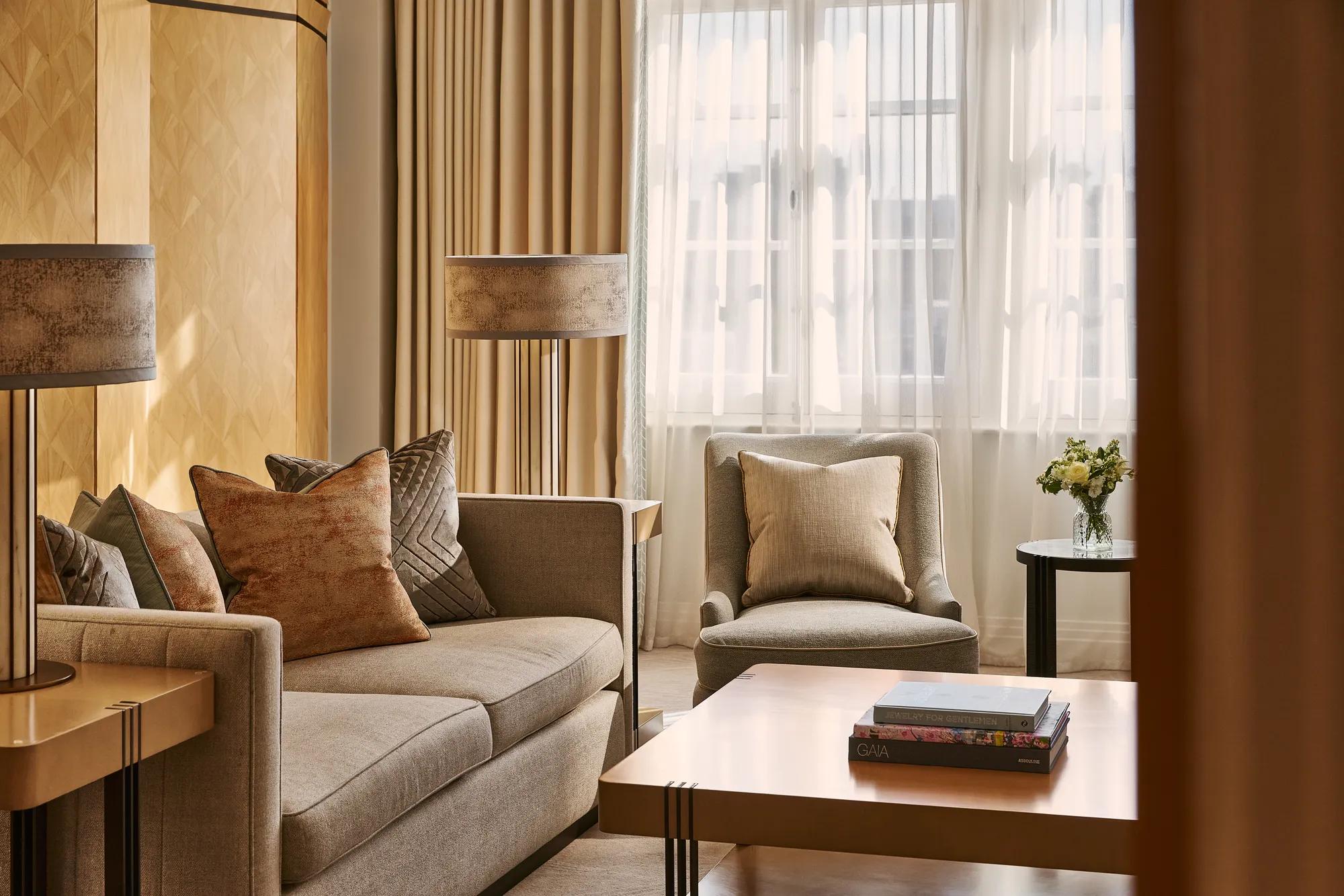Warm-toned sitting area with sofa and armchair, textured cushions, wooden coffee table with books, and window with sheer curtains.