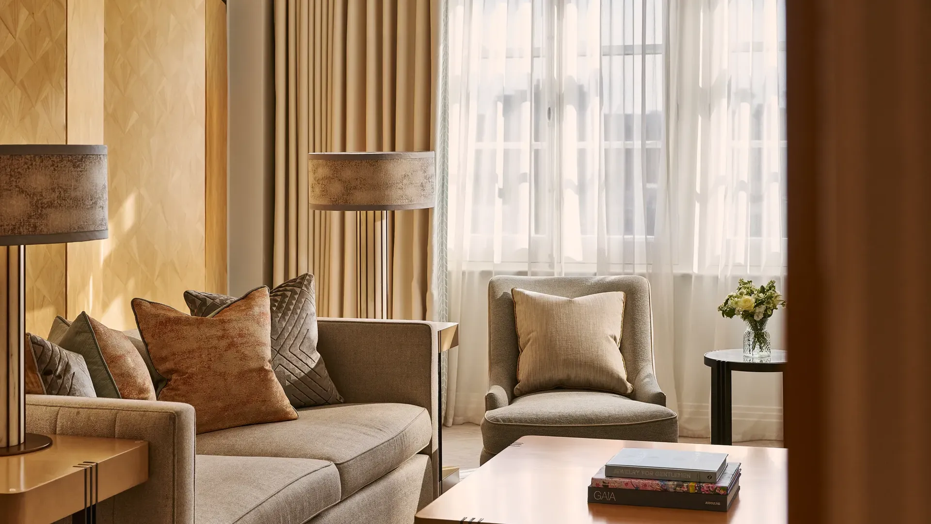 Warm-toned sitting area with sofa and armchair, textured cushions, wooden coffee table with books, and window with sheer curtains.