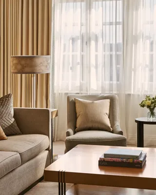 Warm-toned sitting area with sofa and armchair, textured cushions, wooden coffee table with books, and window with sheer curtains.