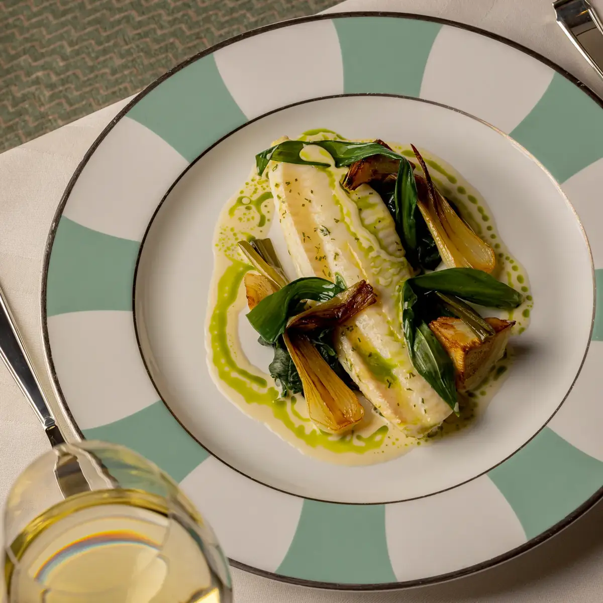 Plated white fish with braised leeks, greens, and herb oil on striped plate, served on white tablecloth with wine glass.