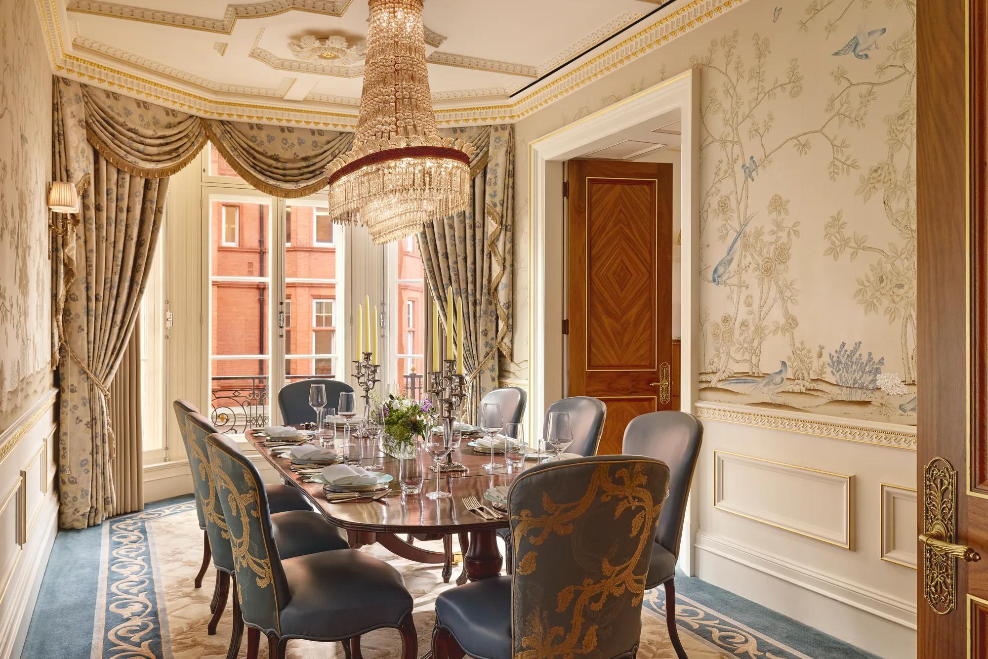 Luxurious dining room with a polished oval table set for eight, upholstered blue and gold chairs, hand-painted wallpaper featuring birds and trees, and a crystal chandelier hanging above.