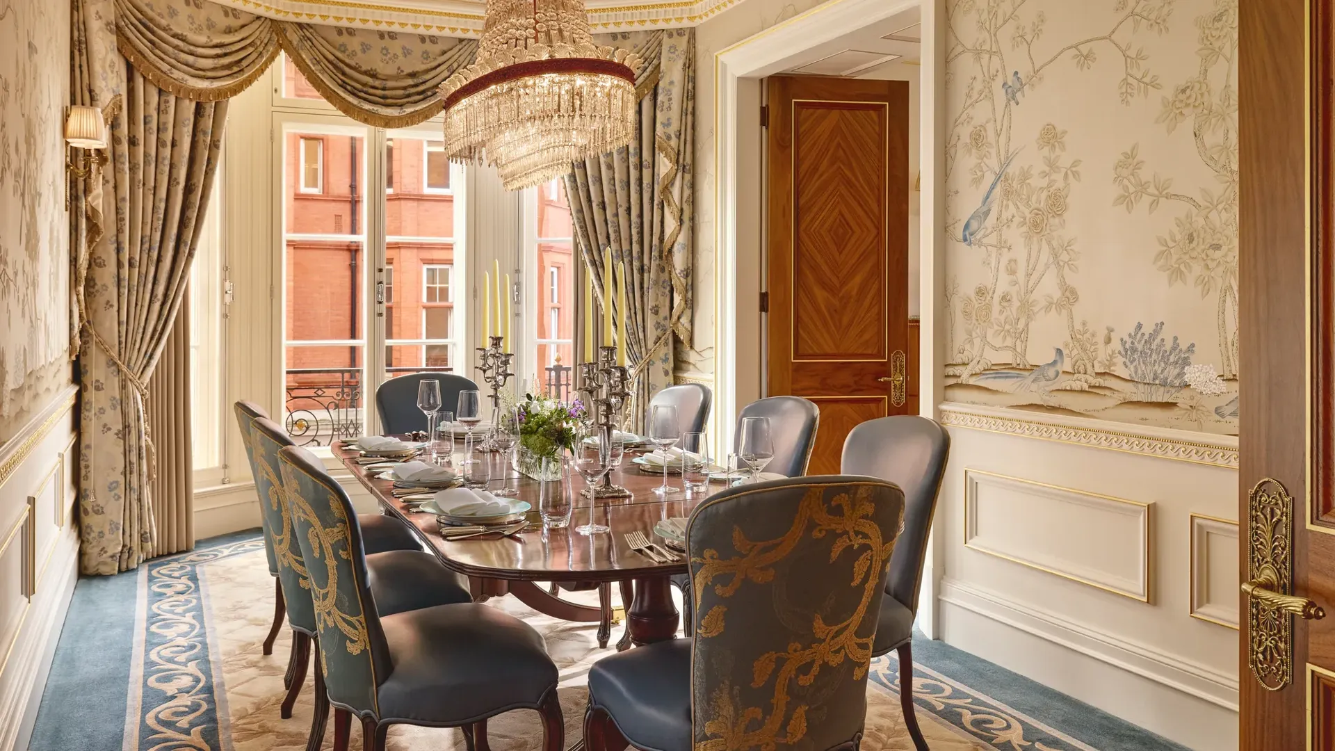 Luxurious dining room with a polished oval table set for eight, upholstered blue and gold chairs, hand-painted wallpaper featuring birds and trees, and a crystal chandelier hanging above.