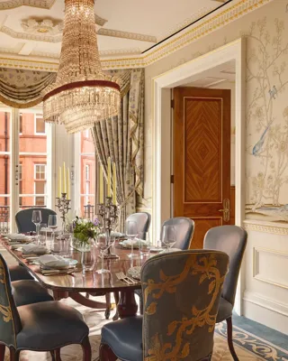 Luxurious dining room with a polished oval table set for eight, upholstered blue and gold chairs, hand-painted wallpaper featuring birds and trees, and a crystal chandelier hanging above.