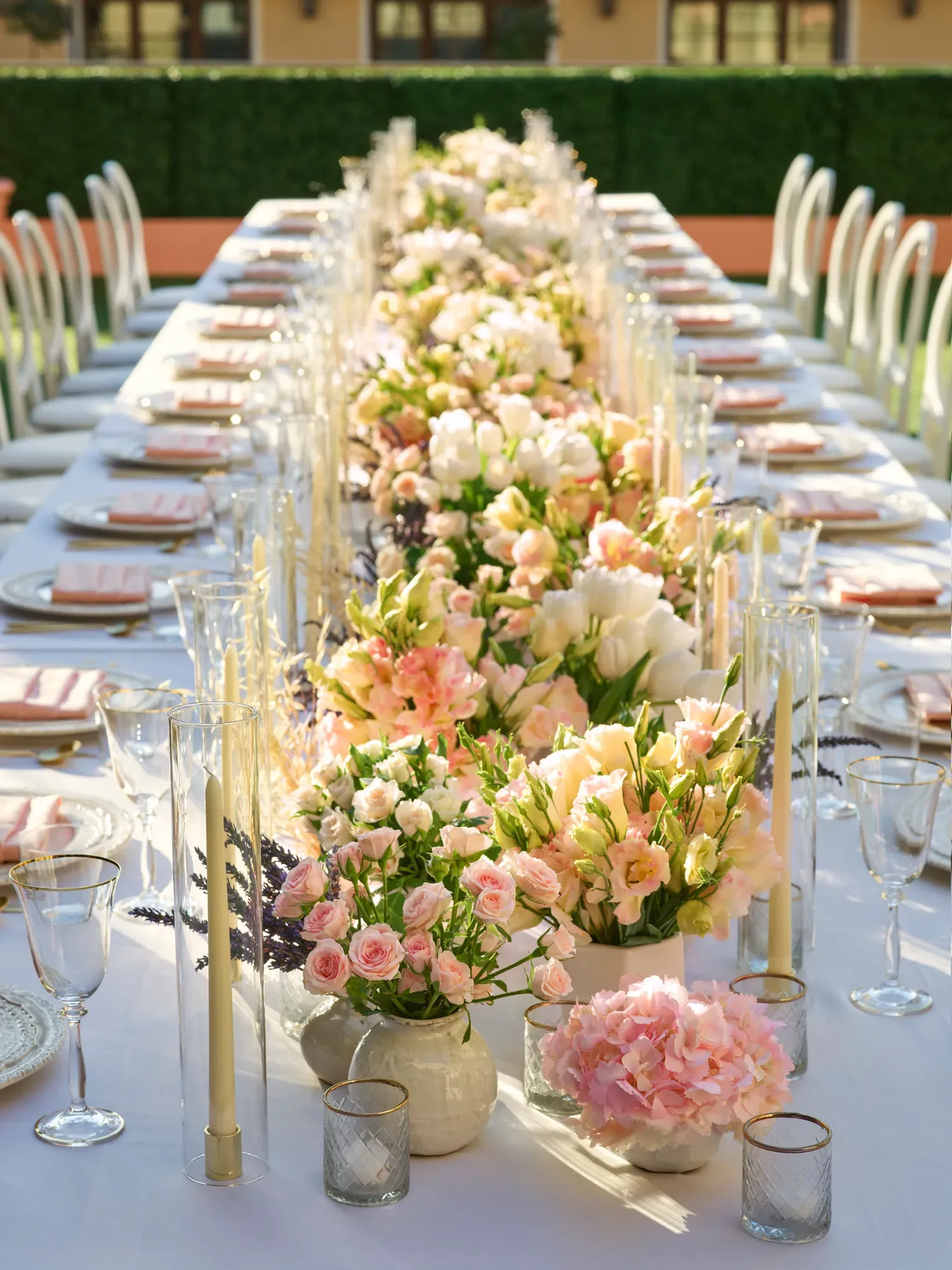 Daytime close-up down the long garden table; lush pastel floral centerpieces and tall clear candle cylinders line white place settings.