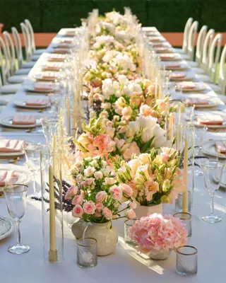 Daytime close-up down the long garden table; lush pastel floral centerpieces and tall clear candle cylinders line white place settings.