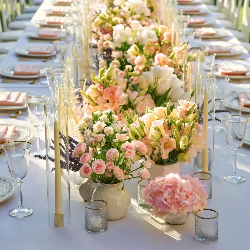 Daytime close-up down the long garden table; lush pastel floral centerpieces and tall clear candle cylinders line white place settings.