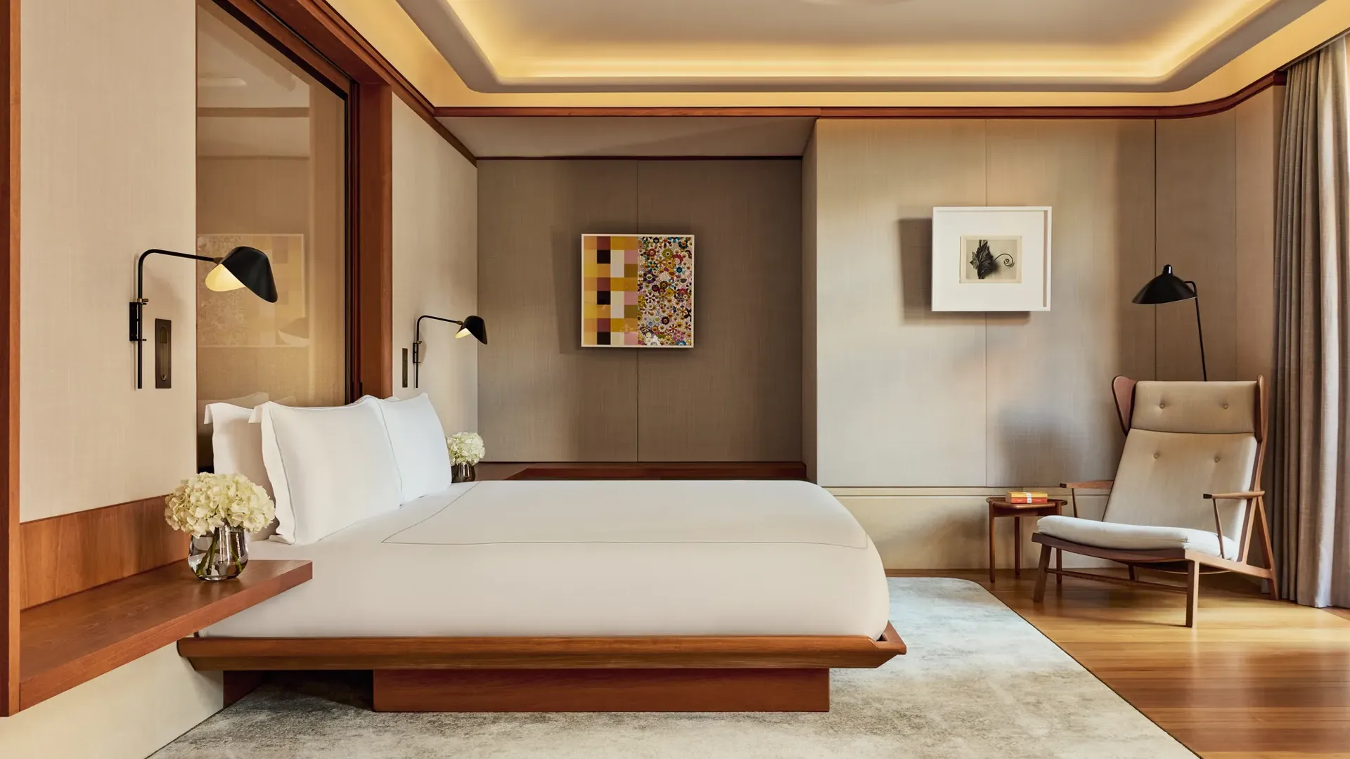 Bright bedroom in the Knightsbridge Suite with a low wooden-framed bed dressed in crisp white linens, wall-mounted reading lights, and mid-century style armchair beneath modern artwork.