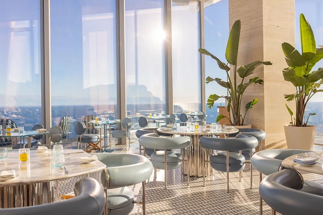 Restaurant lumineux avec tables en marbre, chaises bleues modernes, baies vitrées et vue sur l’océan sous la lumière naturelle.