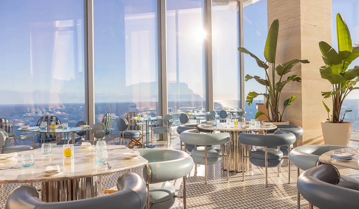 Restaurant lumineux avec tables en marbre, chaises bleues modernes, baies vitrées et vue sur l’océan sous la lumière naturelle.