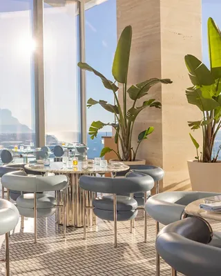 Restaurant lumineux avec tables en marbre, chaises bleues modernes, baies vitrées et vue sur l’océan sous la lumière naturelle.