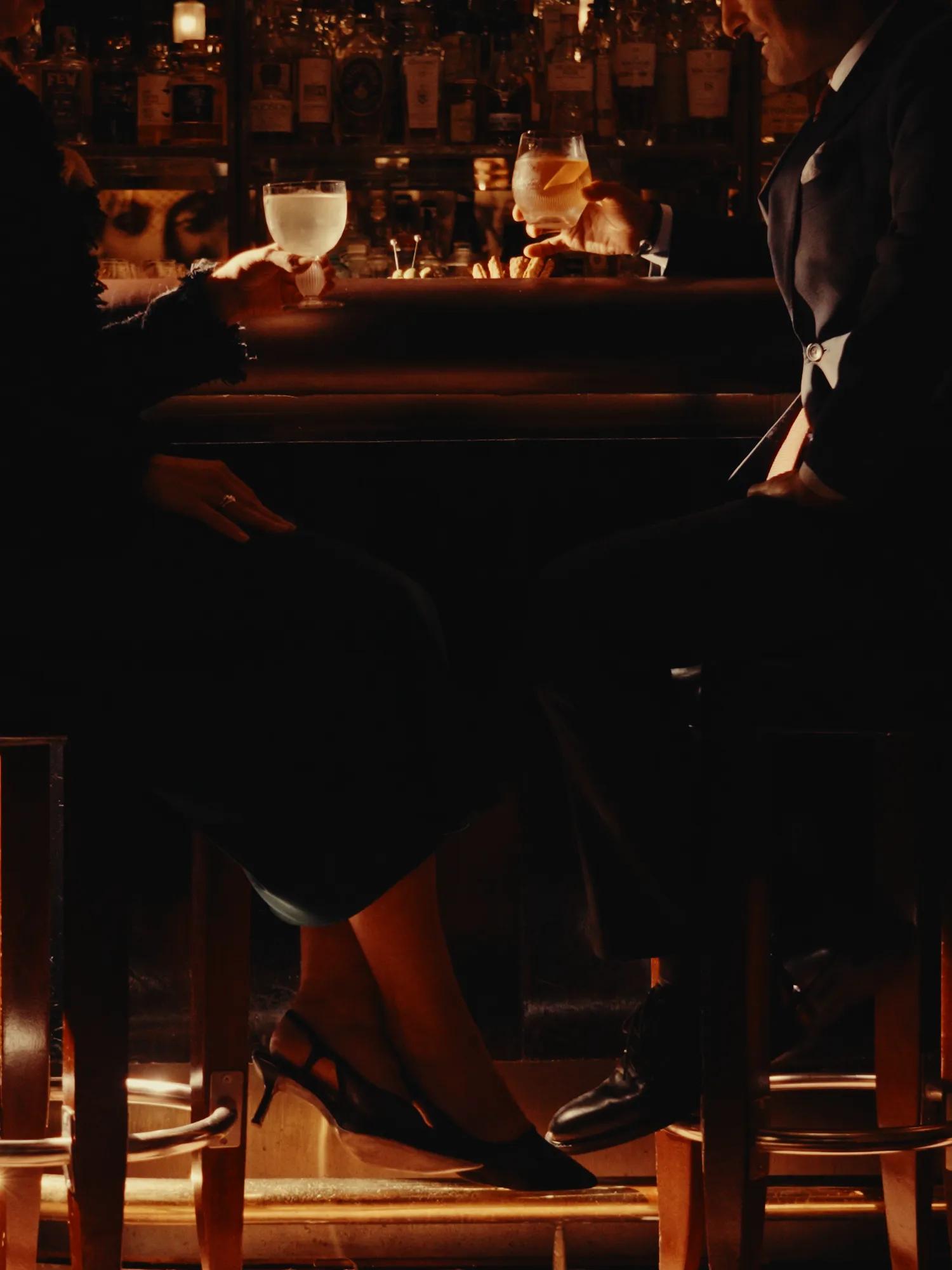 Two guests seated at a softly illuminated bar, enjoying cocktails in an intimate setting framed by warm amber lighting and shelves of spirits.