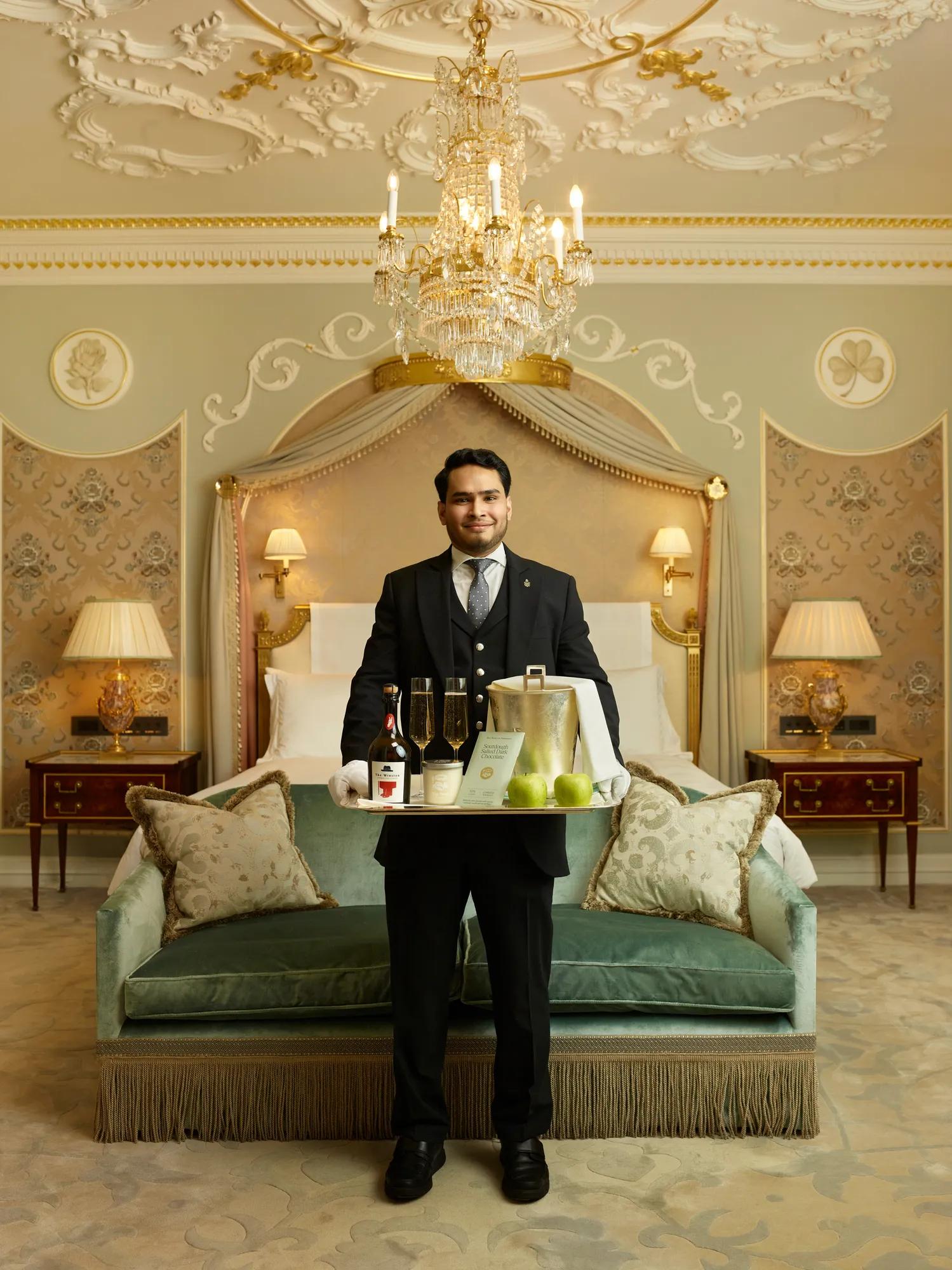 Butler holding welcome tray with cider and apples in luxurious suite beneath chandelier.