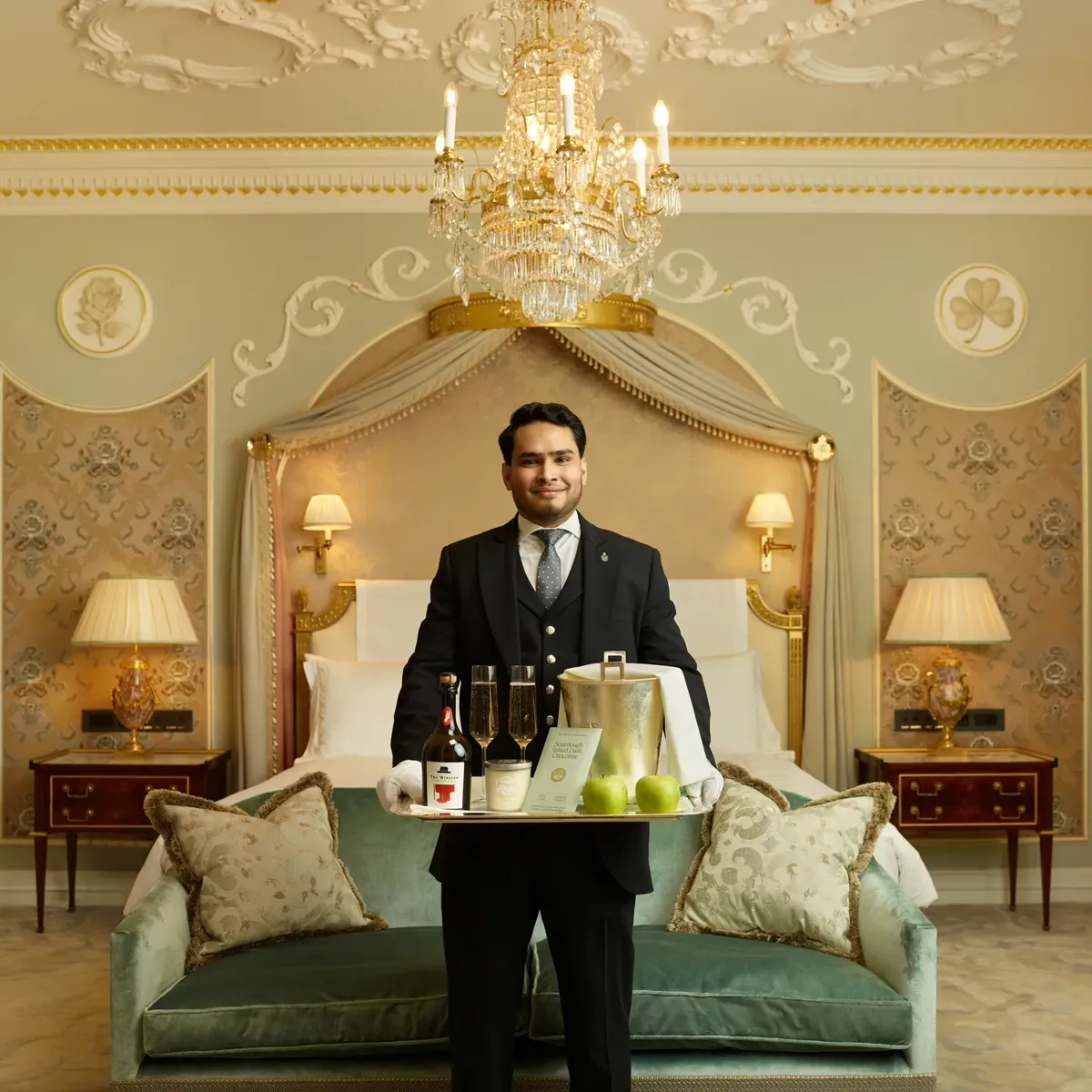 Butler holding welcome tray with cider and apples in luxurious suite beneath chandelier.
