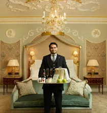 Butler holding welcome tray with cider and apples in luxurious suite beneath chandelier.