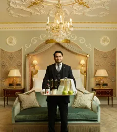 Butler holding welcome tray with cider and apples in luxurious suite beneath chandelier.