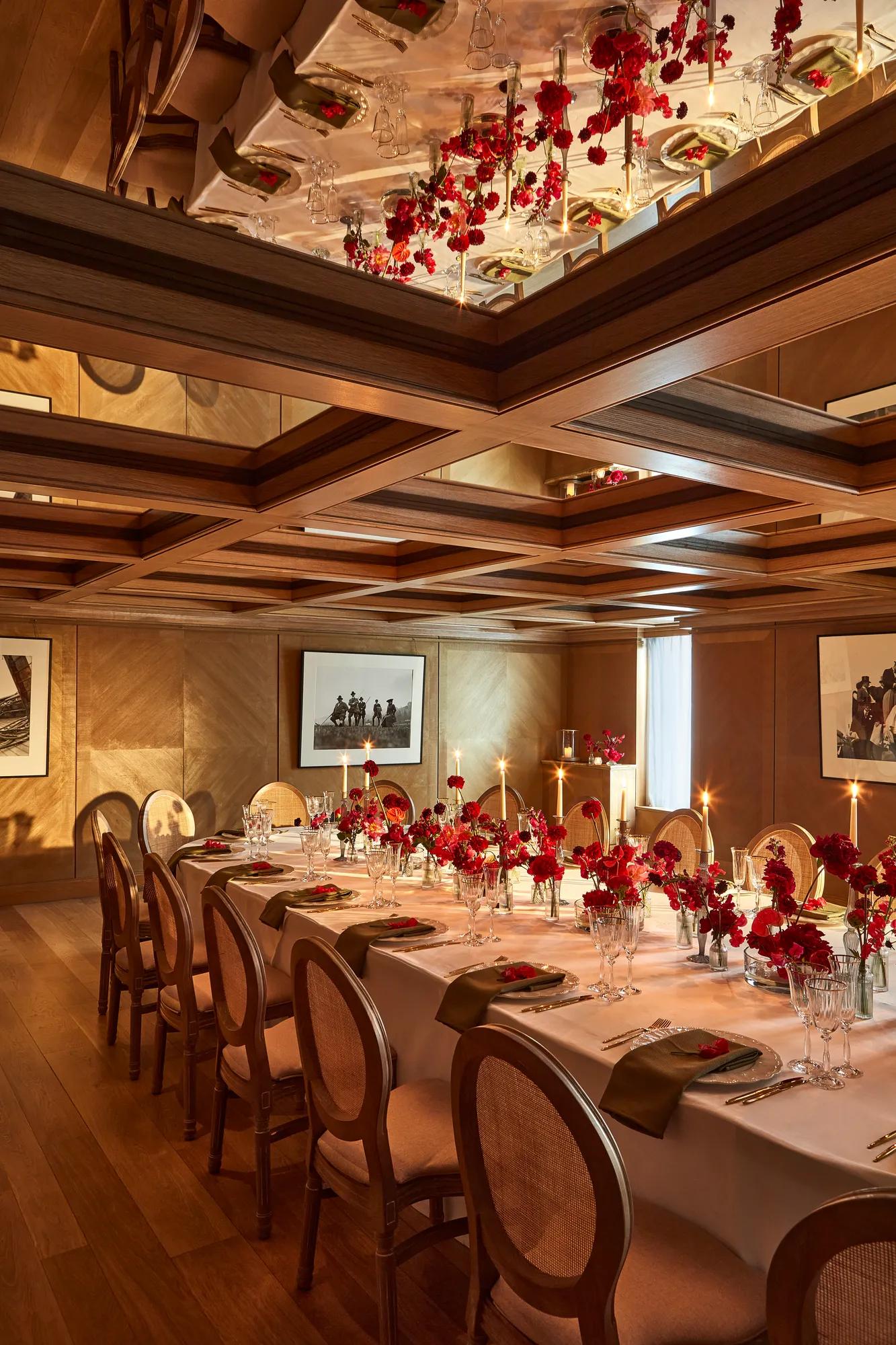 Intimate dining room with a long table set with red floral centerpieces and candles under a mirrored coffered ceiling.