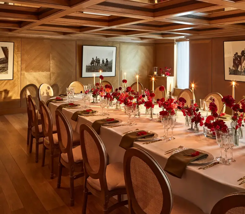 Intimate dining room with a long table set with red floral centerpieces and candles under a mirrored coffered ceiling.