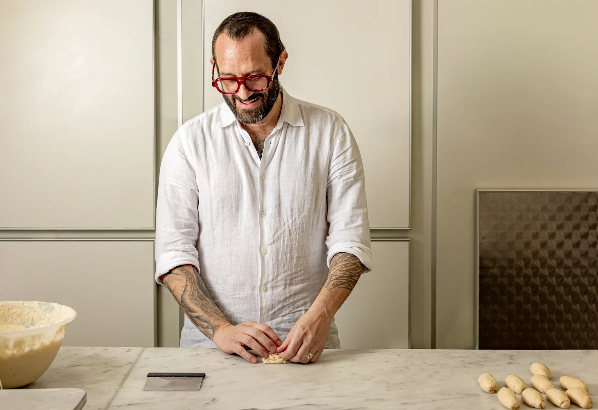 Smiling chef in a white shirt shapes small dough pieces at a marble counter, with a bowl and dough balls nearby