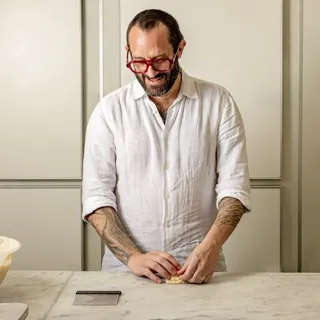 Smiling chef in a white shirt shapes small dough pieces at a marble counter, with a bowl and dough balls nearby