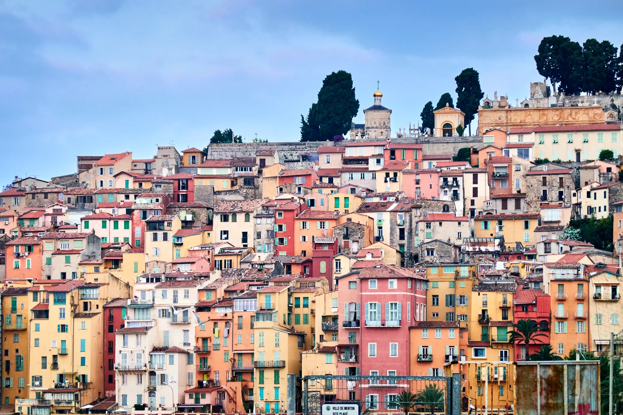 Colourful hillside town with tightly packed pastel buildings, shuttered windows and tiled roofs beneath a blue sky.