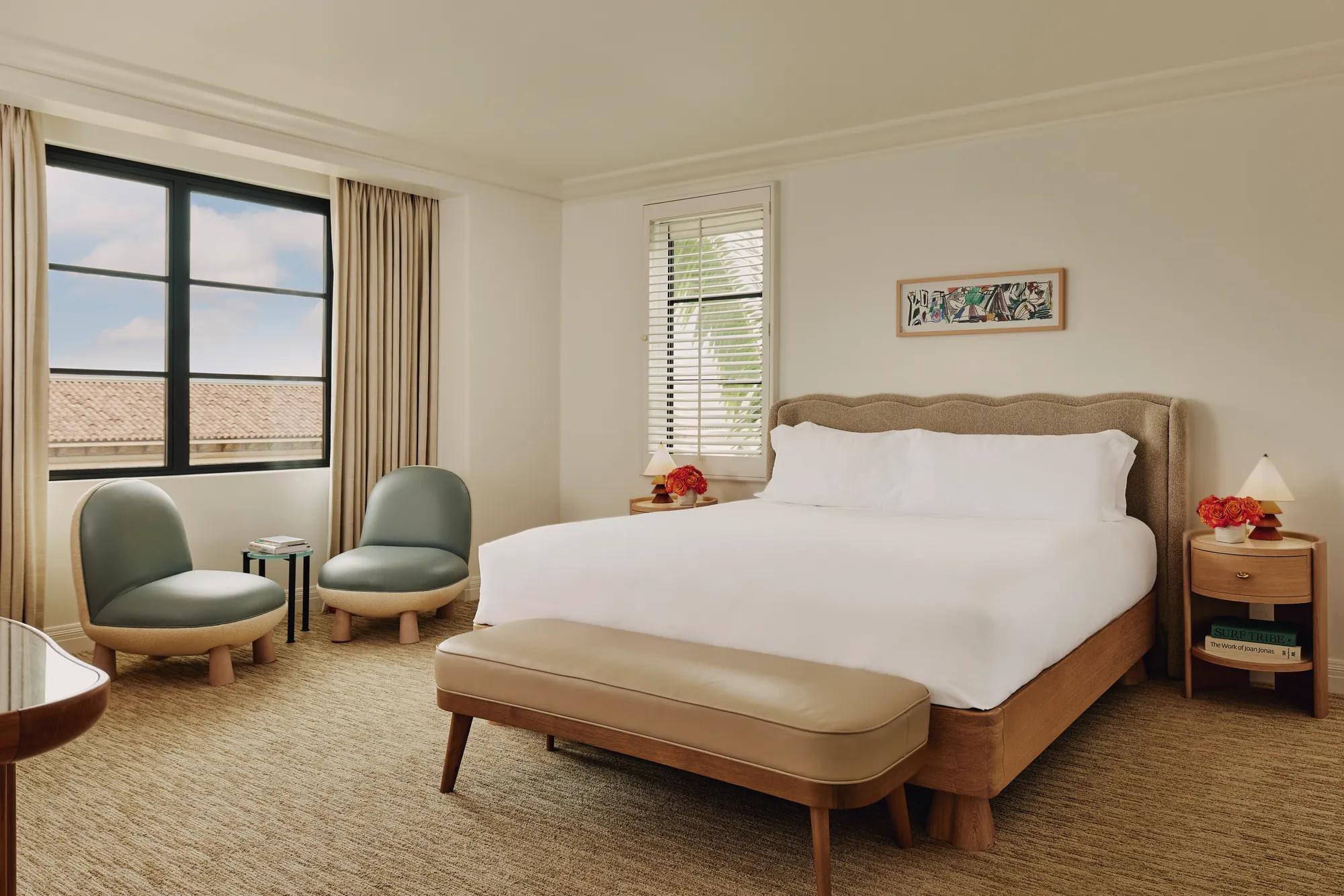 Bedroom with king bed, upholstered headboard, bench at foot, two lounge chairs with side table by the window, and bedside tables with lamps.