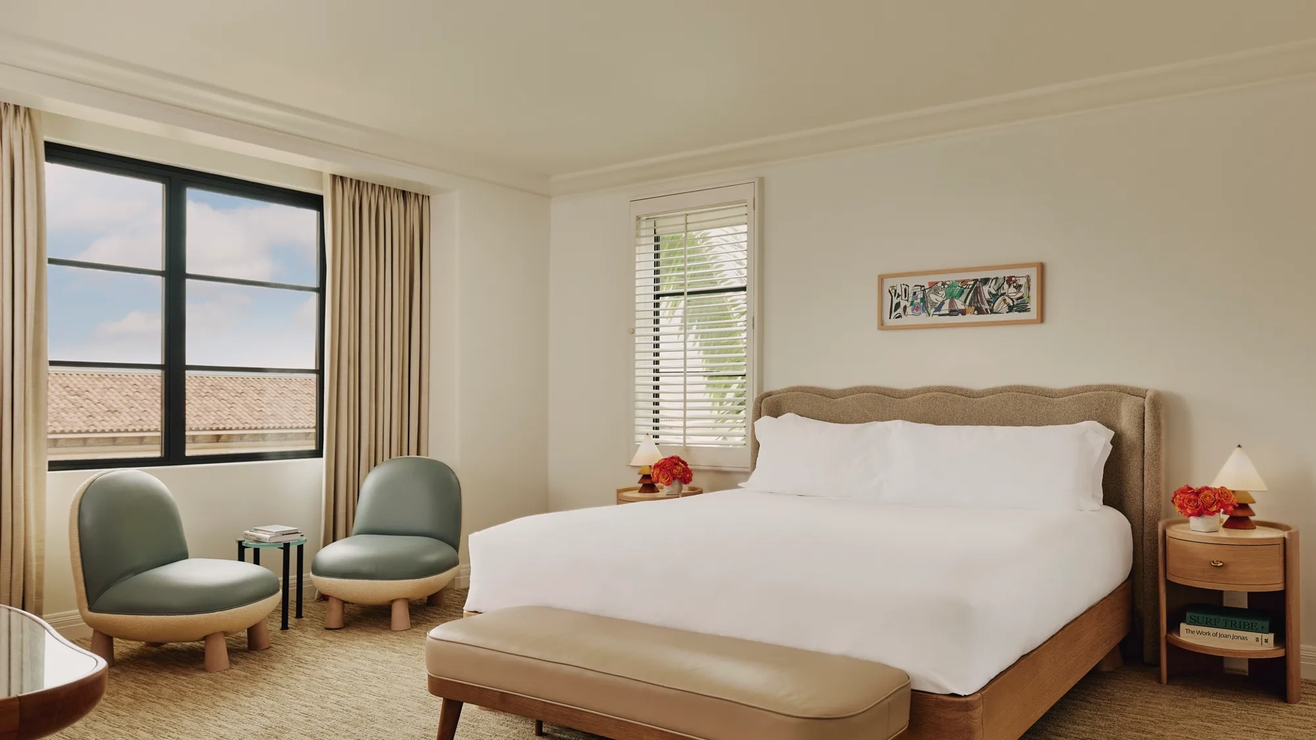 Bedroom with king bed, upholstered headboard, bench at foot, two lounge chairs with side table by the window, and bedside tables with lamps.