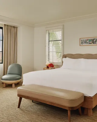 Bedroom with king bed, upholstered headboard, bench at foot, two lounge chairs with side table by the window, and bedside tables with lamps.